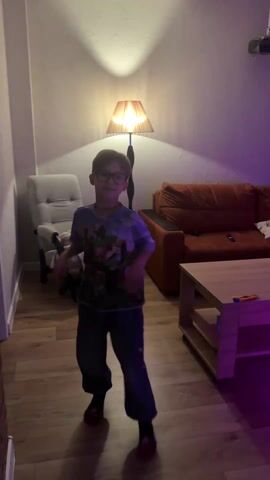 A young boy is dancing in what appears to be a living room. The boy is wearing glasses, a Minecraft-themed t-shirt, blue pants, and red slippers. He is making various arm movements as he dances. The room is illuminated by a standing lamp behind him and a purple light source near the floor. Furnishings in the room include a gray armchair, an orange couch, and a light wood coffee table. A projector is visible mounted on the ceiling in the background. The floor is made of wood.