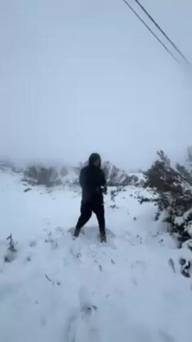 The video shows a person in a snowy environment, throwing snowballs. The person is dressed in dark clothing, including a hooded jacket, and brown shoes. They are standing in a snow-covered area with some bushes and sparse vegetation visible. The weather appears foggy or misty, with limited visibility. The person throws one snowball in the air and then makes another. Power lines can be seen overhead.