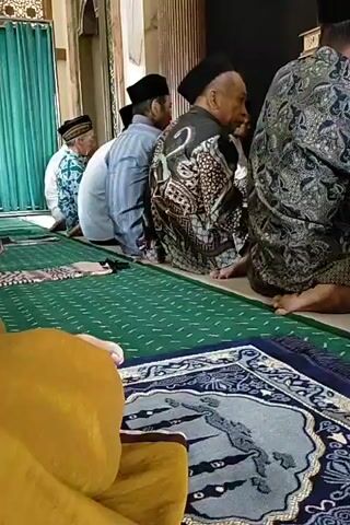 A group of men are seated on the floor, facing away from the camera, in what appears to be a religious setting. They are wearing traditional Islamic attire, including kufis and patterned robes. Two prayer rugs are visible on the floor in front of them. The environment seems to be indoors, with a green curtain visible on the left side of the frame. The men appear to be engaged in prayer or a religious gathering. The lighting is natural, suggesting daylight.