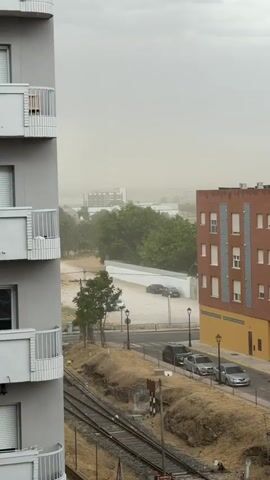 The video shows a view from a window looking out at an urban environment. In the foreground, the side of a gray building with white balconies is visible on the left. To the right, a brick building with an orange base and white window frames is prominent. Below the buildings, a set of train tracks runs horizontally. Beyond the tracks, a street with parked cars is visible, as are lampposts and a small tree. Further in the background, more trees and buildings can be seen, all partially obscured by a thick haze that gives the sky a light brown, dusty appearance. The haziness suggests poor visibility, possibly due to dust or pollution.
