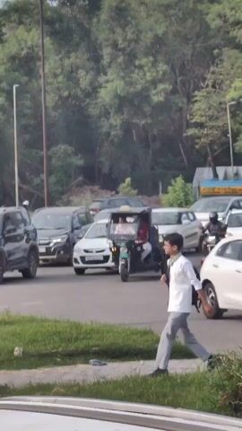 The video shows a street scene with traffic. In the foreground, a young person in a white shirt and grey pants walks across the street, carrying a backpack. In the background, there is a green auto-rickshaw with two people inside, and several cars and motorcycles on the road. The scene appears to be in a city or town, with trees and a pole visible in the background. The lighting suggests it is daytime.