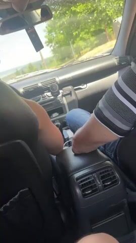 The video shows the interior of a car from a rear passenger's perspective, looking forward. Two individuals are visible in the front seats. The person in the driver's seat is partially visible from the side, with their left arm resting on the center console. The person in the passenger seat is wearing a striped shirt and jeans, and their arm is also resting on the center console, near the gear shift.

The car's dashboard is visible, including the radio, air vents, and a phone mount holding a smartphone. A car air freshener, possibly branded "Areon," hangs from the rearview mirror. The rearview mirror reflects the faces of the two individuals in the front seats.

The view through the windshield reveals a sunny daytime scene. Trees and foliage are visible outside, suggesting a rural or suburban setting. The sky is bright, indicating it is daytime, likely late morning or afternoon given the lighting. The overall mood appears relaxed, with the occupants seemingly engaged in conversation or simply enjoying the drive. There are no distinct audible sounds from the video.

The time stamp "00:00 - 00:04" indicates the short duration of this clip. The location context provided is Zvezditsa, Bulgaria, which aligns with the rural scenery visible. There is text visible on the air freshener.