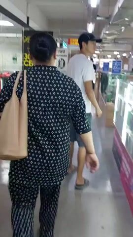 The video shows an indoor marketplace, likely a shopping mall or electronics store, with several stalls and display cases visible. The flooring appears to be concrete. In the foreground, a person wearing a black patterned shirt and pants, carrying a light-colored tote bag over their shoulder, is walking away from the camera. To the right, a person wearing a white t-shirt, dark shorts, and a black baseball cap is walking in the same direction as the first person. Several display cases with glass tops and wooden bases are filled with various items, possibly electronics like phones and accessories. Signs with text in Russian are visible in the background, indicating services like phone registration and repair. The lighting is bright, coming from overhead fixtures. The overall atmosphere seems to be that of a bustling commercial environment.