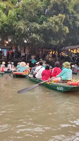 Several boats filled with people are on a body of water. The boats appear to be traditional rowboats, and the people are dressed in casual clothing. Many of the people are wearing conical hats.  The boats are close together, and some have numbers painted on the side. A person in the foreground boat is using an oar to propel the boat. On the bank, a crowd of people can be seen underneath trees. The overall scene suggests a tourist attraction or a recreational area where people enjoy boat rides.