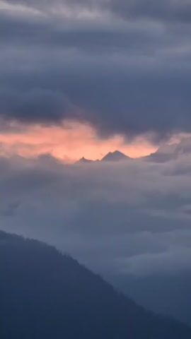 This is a vertical, static shot of a mountainous landscape shrouded in clouds. The sky above is a gradient of dark gray to lighter gray clouds. Peeking through the clouds in the midground, a few mountain peaks are visible, their summits illuminated by a warm, orange glow, suggesting sunrise or sunset. A lower, darker mountain slope dominates the foreground, its texture suggested by the presence of trees, though they are indistinct. The overall atmosphere is serene and somewhat dramatic due to the interplay of light and shadow.