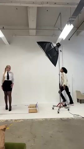 The video shows a photography studio with a white backdrop. Two people are present. One person, appearing to be a model, stands on the left side of the frame, facing the camera. They are wearing a white collared shirt, a black tie, a black skirt, and black stockings. The other person, likely a photographer or assistant, is on the right side of the frame, facing away from the camera and towards the model. They are wearing a cream-colored jacket and dark pants. A large softbox light on a stand is positioned to the right of the model, illuminating the scene. The photographer appears to be giving instructions or adjusting something as they move their hands. The model holds a pose with one hand on their hip. The environment is a professional studio setting, with exposed ceiling beams and industrial lighting fixtures visible.