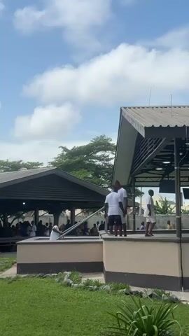 The video shows an outdoor gathering under a covered structure. The sky is blue with scattered white clouds. In the foreground, there is a patch of green grass with some rocks and plants. The covered structure has a metal roof supported by poles. The walls of the structure appear to be made of concrete. There are several people present, most of whom are wearing white clothing. In the middle of the frame, two individuals in white shirts and shorts stand on a raised platform. To the right, another person in white shorts and a white shirt walks away from the camera. Further to the right, a person in a patterned outfit and a person in a pink shirt and blue pants are visible. Inside the covered area, there are screens and tables. On the back wall, there is a sign with text on it, and an air conditioning unit is mounted on the exterior wall. In the background, trees and buildings can be seen. The overall scene suggests an event or service taking place in an open-air venue.