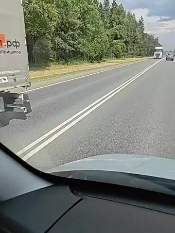 The video shows a view from inside a vehicle driving on a two-lane road. Trees line the right side of the road, and there are some clouds in the sky. A white truck with a blue trailer is seen driving in the opposite lane. Another vehicle is visible further down the road in the distance, also traveling in the opposite direction. The camera perspective suggests the video was recorded by a passenger or from the dashboard of the moving vehicle.