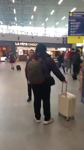 The video shows the interior of an airport terminal. The camera is following a person from behind who is walking with a white suitcase on wheels and a backpack. To the left of this person is another person in a black uniform with a red lanyard. In the background, there are signs with information and flight schedules displayed on large screens. Other travelers are visible in the distance, some with luggage. The environment appears to be well-lit with a modern architectural design.
