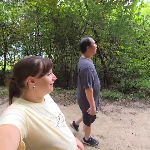 A woman and a man are walking on a dirt path surrounded by trees. The woman is in the foreground, facing left, and is wearing a light-colored t-shirt. She has her hair pulled back and is smiling. The man is behind her and to her right, facing right, and is wearing a dark t-shirt and shorts. He is looking forward and appears to be holding a phone in his right hand. The woman makes hand gestures. The video is shot from a low angle.