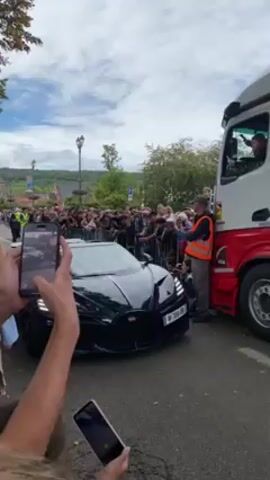 The video shows a dark-colored Bugatti sports car in front of a crowd of people. The car is a convertible, and the top is down. There is a man driving the car, and a young boy in the passenger seat. The car is parked next to a large white truck. The crowd is taking photos and videos of the car. The setting appears to be a town square. The sky is overcast.