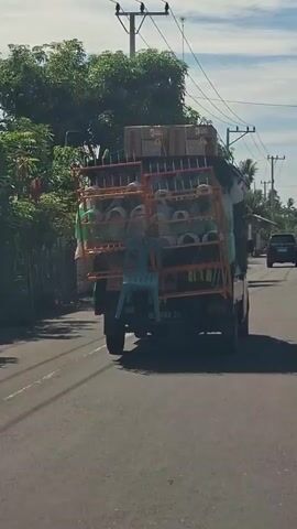 A video captured from behind a truck shows the truck driving on a road. The truck is loaded with several items. On the top of the truck, there are stacked boxes. Below the boxes, there are shelves filled with cylindrical objects, possibly pipes or rolls of material, some of which are white and others are green. A blue plastic chair is attached to the back of the shelves. The truck has a license plate that reads "B 8189 ZG." The road is paved and appears to be in a rural or suburban area, with trees and vegetation visible on the sides of the road. There are power lines and utility poles in the background. The lighting suggests it is daytime.