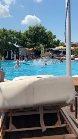 The video shows a sunny day at an outdoor swimming pool. In the foreground, a chaise lounge covered with a light-colored towel is positioned on a wooden deck. The pool is filled with many people, some of whom are swimming or playing in the water, causing splashes. In the background, trees line the perimeter of the pool area, and several yellow umbrellas and structures can be seen, suggesting a resort or lounge area. The sky is blue with scattered white clouds.