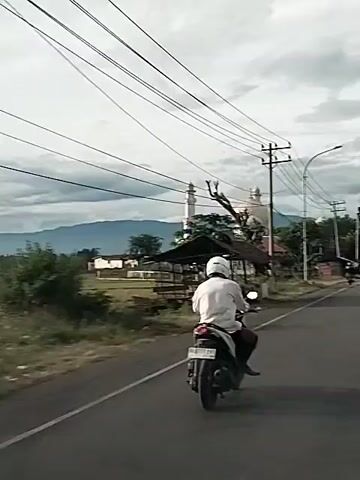 A motorcycle rider wearing a white helmet and white shirt is traveling on a paved road. The rider is in the foreground, facing away from the camera. The motorcycle has a license plate that is partially visible. To the left of the road is a field and a small wooden structure. In the background, there are buildings, trees, power lines, and mountains. The sky is cloudy. There are other vehicles on the road, including a white van and a bus. The video is short and shows the rider traveling forward.
