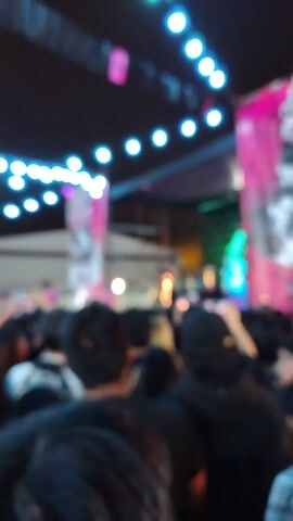 The video shows a performance on an outdoor stage at night. A band plays on a stage decorated with pink banners featuring stylized imagery. The scene is lit by a string of blue and purple lights hung above the crowd. People in the audience are holding up their phones to record the performance, and several hands are raised in the air. A sign reading "VEN A VER LAS MARCAS MAS COOL" is visible above the crowd near the edge of a building.