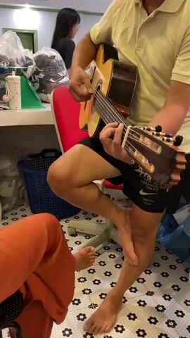 A man in a yellow polo shirt and black shorts sits on a red chair, playing an acoustic guitar. His left leg is crossed over his right, with his left foot resting on the floor. A person wearing orange pants sits on the left side of the frame, with their feet also visible on the floor. The room has a tiled floor with a black and white floral pattern. In the background, there are shelves with various items, including clothes and bags, and a blue laundry basket. A person with long dark hair is partially visible in the background, standing near a green object.