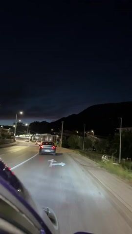 This video shows a rear-facing view from inside a vehicle driving at night on a road. The road is paved and has visible lane markings, including directional arrows illuminated by the vehicle's headlights. Several other cars are driving on the road, with their taillights visible. Streetlights illuminate parts of the scene. On the right side of the road, there is a sidewalk, vegetation, and buildings. In the distance, there are hills or mountains silhouetted against a dark sky. The sky shows a gradient of dark blue to lighter hues near the horizon, with some clouds. The overall impression is of a suburban or semi-urban area at dusk or night.
