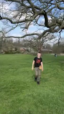 The video depicts a young person walking across a grassy field toward the camera. The person wears glasses, a black t-shirt, and khaki pants, and carries a small, tan-colored bag over their shoulder. Behind the person, there is a large, bare tree with many branches against a backdrop of a bright sky with light clouds. In the distance, other figures can be seen walking across the grass. The person walks up to the camera and says "Hello."