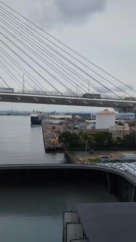 The video shows a view from a ship looking towards a large cable-stayed bridge over a body of water. The bridge has multiple lanes of traffic, with trucks visible on the roadway. The water appears to be a harbor or river, with docks and industrial buildings on the shore. A large ship is moored at one of the docks. The sky is overcast, suggesting cloudy weather. The video pans slightly from right to left, revealing more of the bridge's structure and the surrounding port. The lower portion of the video shows the deck of the ship from which the footage is being recorded.