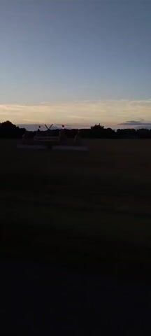 The video opens with a wide shot of a rural landscape at dusk. In the center of the frame, a castle-like structure made of hay bales is visible in a field. The structure has multiple towers, flags, and a banner that reads "GRASS CASTLE HERE!". The sky is a pale blue with streaks of orange and pink from the setting sun. As the video progresses, the camera pans to the right, revealing more of the field and trees in the distance. The sun is low on the horizon, casting long shadows. The final shots show the sun fully set, with the sky transitioning to a deeper blue. The video appears to have been filmed from a moving vehicle, as indicated by the slight blur and the perspective. There are no people visible in the video.