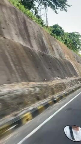 The video shows a view from the perspective of someone riding a motorcycle on a road with a retaining wall on the left. The retaining wall is made of concrete and appears to have some erosion and cracks, with debris visible at its base. There are some trees and vegetation growing on top of the retaining wall. The road itself is asphalt with yellow and black striped markings along the edge. Several cars are driving on the road ahead. The sky is visible in the background, and it appears to be daytime with a clear or partly cloudy sky. In the distance, there are more trees, palm trees, street lights, and buildings. The overall environment seems to be a suburban or semi-rural area with a focus on infrastructure and transportation.