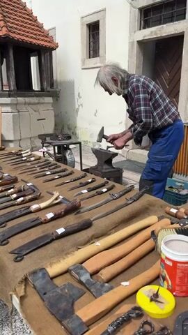 An elderly man with long, light gray hair, wearing a plaid shirt and blue work pants, is actively engaged in blacksmithing outdoors. He is shown hammering a small metal piece on an anvil, which has the number "30" visible on its side. He appears focused and skilled in his craft.

In the foreground, a large table covered with a brown cloth displays a wide assortment of handcrafted metal items. These include numerous knives, daggers, and axes, many featuring detailed handles made of wood or twisted metal. Other tools and brushes are also on display, suggesting a stall at a market or a craft fair. A white container and a yellow container are also visible on the table.

The setting is outdoors on a bright, sunny day. In the background, there's a traditional white building with windows and a wooden door, indicative of old European architecture. To the left, a structure with a red-tiled roof, possibly a well, adds to the historical ambiance. The scene depicts a demonstration of traditional craftsmanship, likely in Častá, Slovakia.