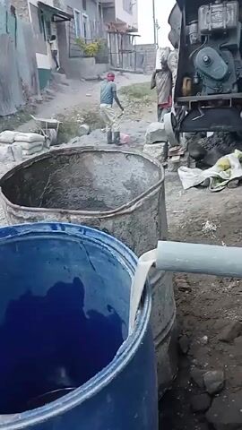 The video shows a construction site. A blue plastic barrel in the foreground is being filled by a grey pipe. Behind it is a rusted metal barrel. A man carries a white container, presumably filled with materials for the construction. He walks past a cement mixer, which is not currently in operation. Another man is working in the background, possibly mixing materials on the ground. The site is dusty, with piles of rocks, bricks, and building materials scattered around. A few buildings are visible in the background, suggesting the site is located in a residential area.