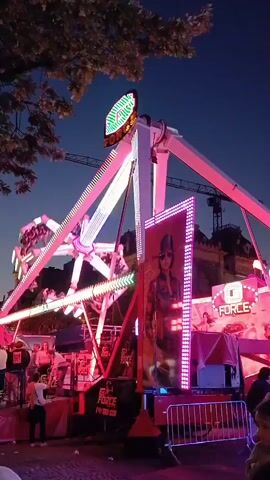 A fairground ride called "The Force" is in operation. The ride consists of a central vertical tower with two arms extending outward and upward. Each arm has a gondola at its end. The gondolas are populated with people. The ride is illuminated with numerous bright pink and green lights, giving it a vibrant appearance against the twilight sky. A large banner featuring a person wearing sunglasses and a helmet is displayed on the side of the ride's base. People are gathered around the ride, some appearing to be waiting in line. The ground is paved with cobblestones. Trees with foliage are visible in the upper left corner of the frame.