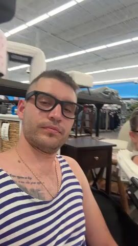 A man and woman are sitting in chairs in a store. The man is in the foreground, wearing a blue and white striped tank top and glasses. The woman is in the background, wearing a gray tank top and orange sunglasses. She is holding up two fingers in a peace sign. The store appears to be a home goods store, with shelves of merchandise and furniture visible in the background. A sign above the shelves reads "Decorate in style for less."
