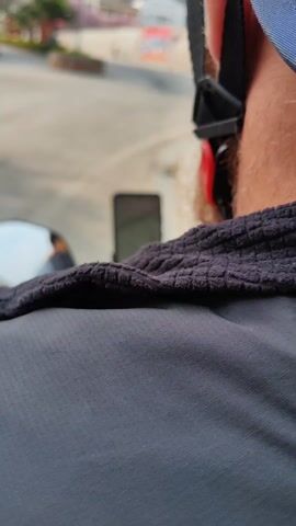 From a rear-view perspective, the video shows a person wearing a helmet and a dark gray textured jacket on a scooter. The person is consulting a smartphone mounted on the scooter's handlebars displaying a map application, possibly for navigation. Their hands are visible as they interact with the screen. The scooter is on a paved road, and another scooter passes by. The background includes buildings, trees, and a clear sky.