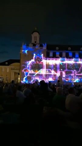 The video captures an outdoor nighttime event featuring a building illuminated with a vibrant light show. The building's facade is adorned with abstract, geometric patterns and colorful projections, primarily in shades of blue, purple, pink, and yellow. Beams of light emanate from above, casting rays into the dark sky. In the foreground, a crowd of people is seated on the ground, facing the illuminated building, appearing to watch the spectacle. The atmosphere suggests a public celebration or festival.