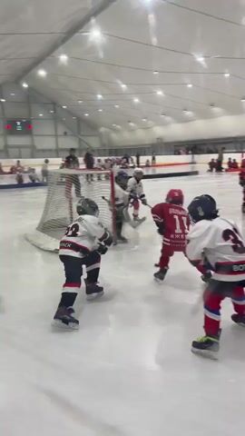 The video shows a hockey game played by young children indoors. The ice rink is brightly lit by overhead lights, and there are spectators in the background behind a barrier. In the foreground, a goalie in full gear stands in front of a net. Several young players in colorful uniforms are skating on the ice, some with hockey sticks. One player in a white jersey with the number 31 on the back is prominent in the frame, facing away from the camera and towards the action. Another player in a red jersey with the number 11 is visible. The game appears to be in progress, with players positioned around the net and in the center of the rink.