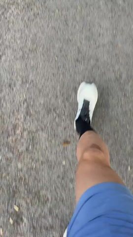 The video shows a first-person perspective of someone walking on a textured gray surface, likely pavement. The person is wearing blue shorts and white and black athletic shoes. The camera angle is from the perspective of the walker, looking down at their legs and feet as they move forward. The background is blurred, indicating motion.