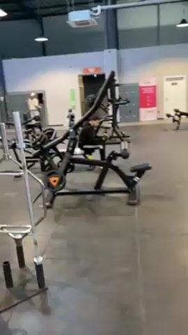 The video shows a wide shot of the interior of a gym. The gym has a high ceiling with exposed metal beams and multiple large windows letting in natural light. There are various weight machines and benches arranged throughout the space, predominantly in black and orange. People can be seen scattered around the gym, presumably working out, though their specific activities are unclear due to the distance. One person, wearing a brightly colored shirt and a hat, walks toward the camera, carrying a white object that may be a towel or bag. The camera pans slightly to showcase the size and layout of the gym.