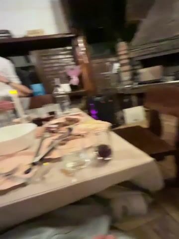 The video shows a blurred, slightly shaky scene from a dining event, possibly a party or celebration. A long table dominates the foreground, covered with a white tablecloth and set with plates, cutlery, and glasses. Food items are scattered across the plates. Several people, all wearing white shirts, are standing around the table. One person is visible holding a smartphone, possibly recording or taking pictures. The environment appears to be indoors, with wooden beams or structures visible in the background. The overall impression is one of a gathering with people engaged in dining and socializing.