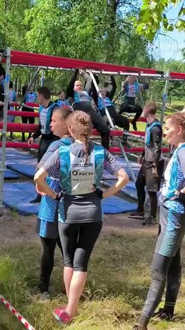 The video shows a group of people participating in an outdoor obstacle course. The course appears to be a series of monkey bars suspended from a metal structure. Several participants are seen on the monkey bars, either in the process of climbing across or waiting their turn. They are wearing athletic clothing, including vests with numbers and logos. In the foreground, two women are standing and observing the obstacle course. The environment is outdoors with trees and grass visible, suggesting a park or wooded area. The lighting indicates it is daytime.