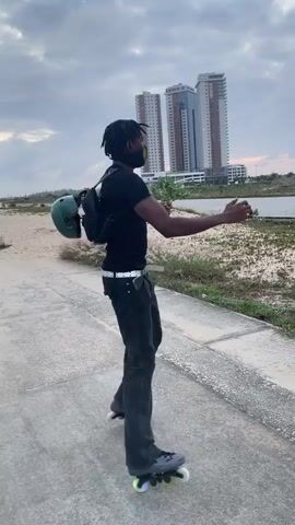 A person wearing black clothing and rollerblades skates on a paved path. The person is also wearing a black mask and a backpack. In the background, there are buildings and a cloudy sky.