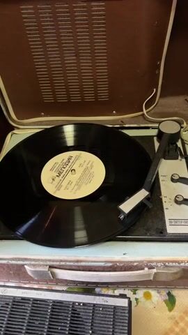 A vintage suitcase record player is open with a black vinyl record playing on its turntable. The record has a white label with text in a foreign language. The record player is brown and light blue. A needle is resting in the groove of the record. The record player is sitting on a surface with a floral pattern.
