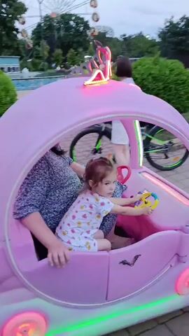 A child is in the driver's seat of a pink toy car that resembles a carriage, holding a steering wheel. The car has a heart-shaped light on the back and lights on the front and sides. The car is on a paved walkway in a park setting. In the background, there is a woman standing next to a bicycle, a Ferris wheel, some trees, and buildings. The sky is cloudy. The video pans to the right, following the toy car as it moves away.