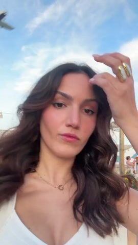 The video begins with a close-up of a woman with long, wavy brown hair, wearing a white top and a delicate necklace. She is looking towards the camera with a slight smirk and adjusting her hair with her right hand, which is adorned with a large ring. The background is slightly blurred but shows a bright sky with scattered clouds and what appears to be an outdoor setting with people and structures.

The camera then pans upwards and to the right. The focus shifts to a large, colorful beach umbrella with orange, white, and teal panels. A small airplane is visible flying in the distance against the blue sky. Below the umbrella, a blonde woman in a red top is visible, as well as other indistinct figures and what looks like some string lights. The overall impression is that of a lively outdoor gathering, possibly at a beach or a resort, during daylight.
