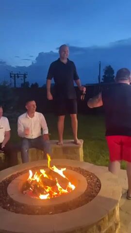Four men are gathered around a fire pit outdoors at dusk. A man in a black polo shirt and shorts is standing on the edge of the fire pit, holding a bottle and appears to be dancing or performing. Another man in a white dress shirt is sitting to the left of the fire pit, looking at the man standing. A man in a black t-shirt and red shorts is standing to the right of the fire pit, facing away from the camera. Another man is partially visible on the far left, wearing a white t-shirt. The sky is dark blue with some clouds. The fire pit is lit with a bright orange flame.