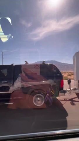 The video shows a dark colored Dodge Ram 2500 truck driving on a highway.  The truck is moving to the right of the frame. The background shows a desert landscape, mountains in the distance, power lines, and a blue sky with scattered clouds. The sun is shining brightly.