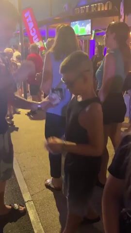 The video shows a group of people gathered outdoors at dusk. A young boy in a black tank top and shorts is dancing in the foreground, moving his arms and head to the music. He is wearing sunglasses and has visible tan lines. To his right, a young girl in a black t-shirt with a tropical graphic is looking at him. In the background, other people are visible, some holding drinks. There is a banner that says "DUCATI" and a screen displaying what appears to be a sporting event. The environment looks like an outdoor market or street festival.