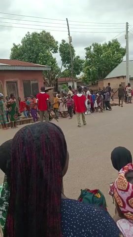 The video shows a group of people walking down a paved street in a village. The street is lined with low, simple buildings and trees. A utility pole with wires runs alongside the road. The sky is overcast. Most of the people are wearing casual clothing, and several individuals are wearing red capes. They seem to be walking together in a procession or parade. The camera is held by a person who is part of the crowd, focusing on the movement along the street. Several people, including children, are visible in the foreground, watching the procession.