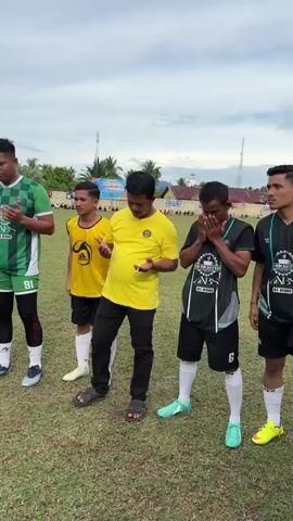 The video shows a group of people, mostly young men in soccer uniforms, standing on a grassy field. They appear to be gathered for prayer, with several individuals with their hands clasped together in front of them. Some of the men are wearing green and white soccer jerseys, while others are wearing black and green jerseys. One person is wearing a bright yellow shirt and black pants. In the background, there are trees, buildings, and what looks like a soccer field with spectators. The sky is overcast. The camera pans slightly, showing different angles of the group.