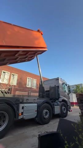 A dump truck with an orange bed is shown from a low angle. The bed of the truck is tilted upwards, dumping a load of light brown sand onto the ground. The truck has multiple axles and large tires. In the background, a brick building with windows and scaffolding is visible, as well as a clear blue sky. The ground appears to be a mixture of dirt and concrete. A few dark green plants are in the foreground, partially obscuring the bottom of the frame. The overall scene suggests a construction or landscaping site.