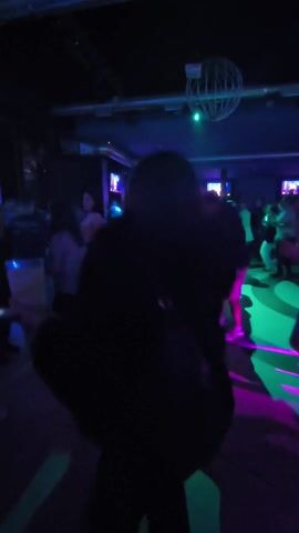 The video shows a person dancing at a crowded dance club or party. The lighting is dark, with colored spotlights creating a dynamic, multi-colored effect. The person is wearing a black jacket over a white shirt and is facing the camera while smiling. They are moving their arms and body to the music. Other people are dancing in the background. The scene includes a disco ball and television screens.