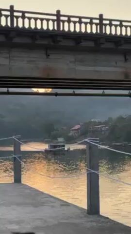 The video shows a lake scene viewed from under a bridge at sunset. The sun’s reflection shimmers on the water's surface. In the middle of the lake, a small house-like structure floats. On the shore, some houses are visible amidst trees on a hill. A concrete pier with a rope railing is in the foreground.