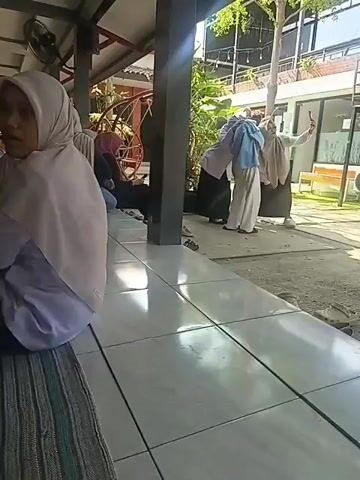 The video shows an outdoor seating area with several people. In the foreground, the back of a person wearing a white hijab and a purple garment is visible. To the left, a person in a pink hijab and black attire is seated. In the mid-ground, three individuals are standing and interacting. One person in a light blue hijab and white pants is holding a phone and appears to be filming or taking a picture. Another person in a beige hijab and black skirt is standing next to them, also looking at a phone. A third person in a white top and black skirt and hijab is standing further to the right, holding a phone and looking towards the other two. The background shows a building with outdoor seating, trees, and some greenery. The lighting suggests it is daytime.