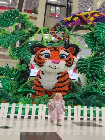 A young girl with blonde hair runs towards the camera. She is wearing a light-colored dress with a floral pattern and appears to be in motion. Behind her, a large, colorful balloon sculpture depicts a tiger sitting amidst jungle-themed balloon decorations, including trees and foliage. The setting appears to be indoors, possibly a shopping mall or public space, with a tiled floor visible. The tiger sculpture is a prominent feature of the scene, with the girl running in front of it. The video captures a playful moment with the child interacting with the elaborate balloon art.